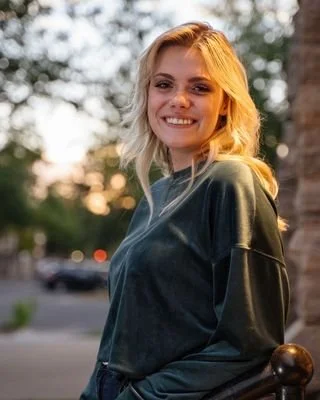 A smiling young woman with blonde hair in a black jacket outdoors at sunset.