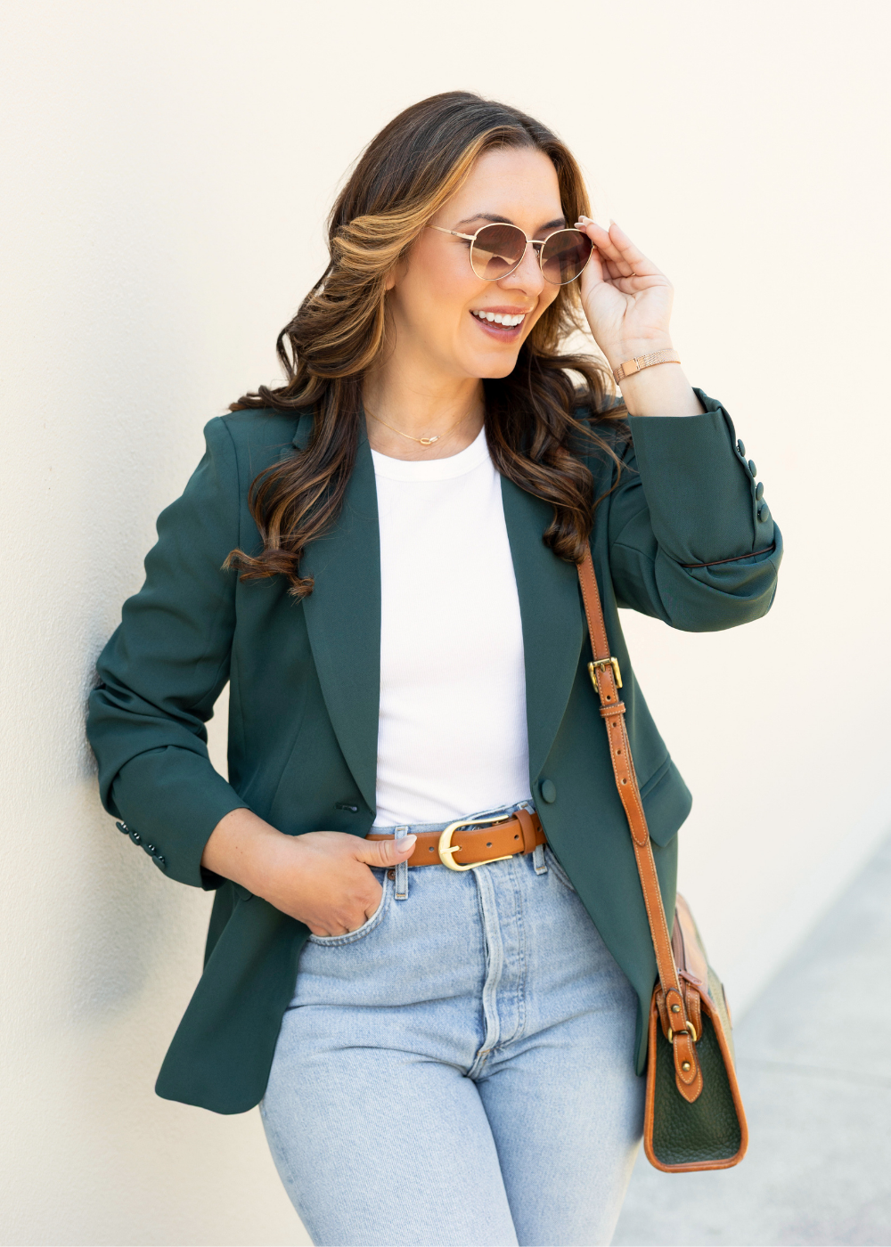 Samantha Rae wearing sunglasses, a green blazer, white t-shirt, light blue jeans, and a brown belt, smiling and standing against a white wall.