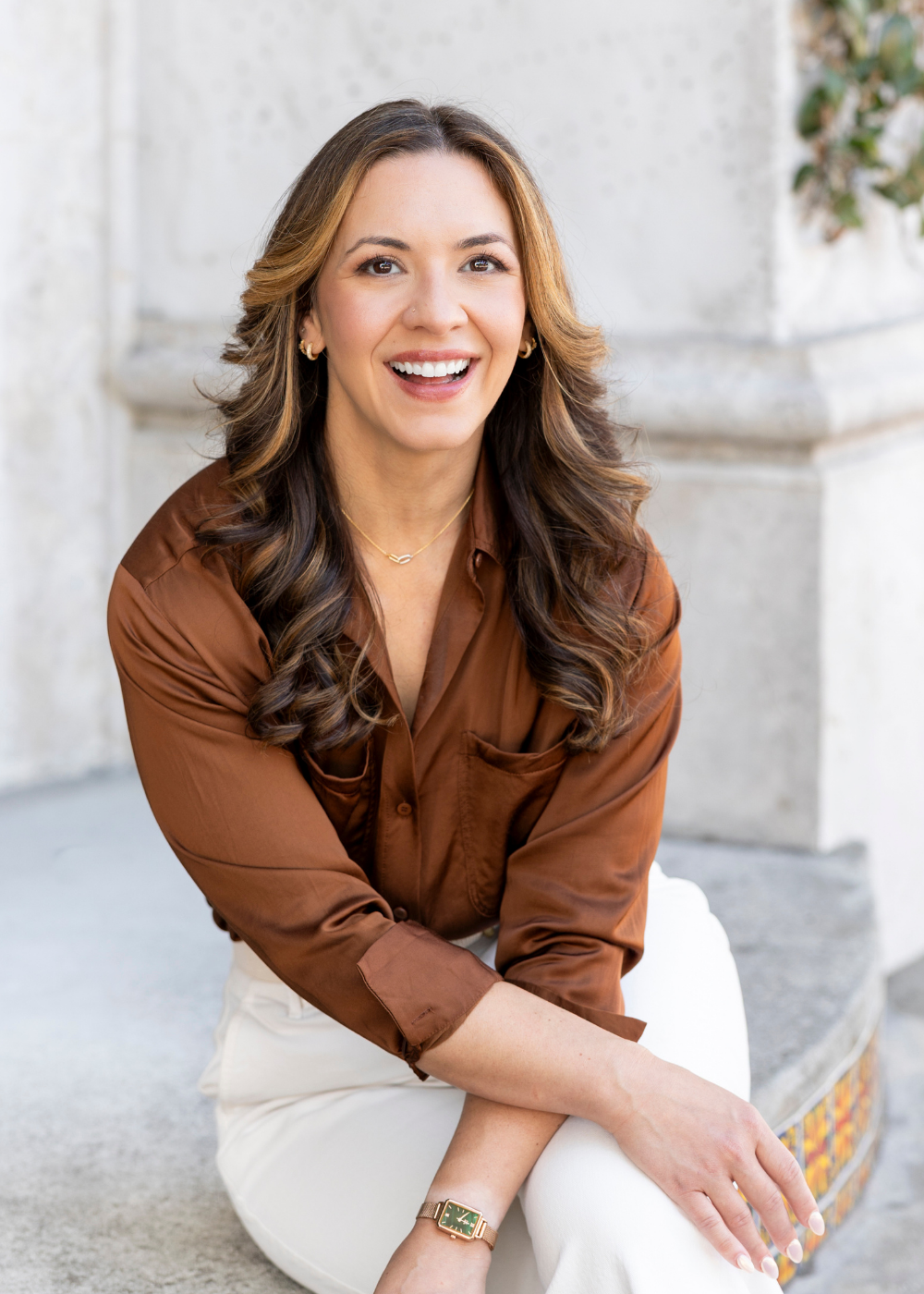 Samantha Rae, a licensed psychologist, smiling, wearing a brown blouse and white pants, sitting outdoors on steps with a concrete wall background.