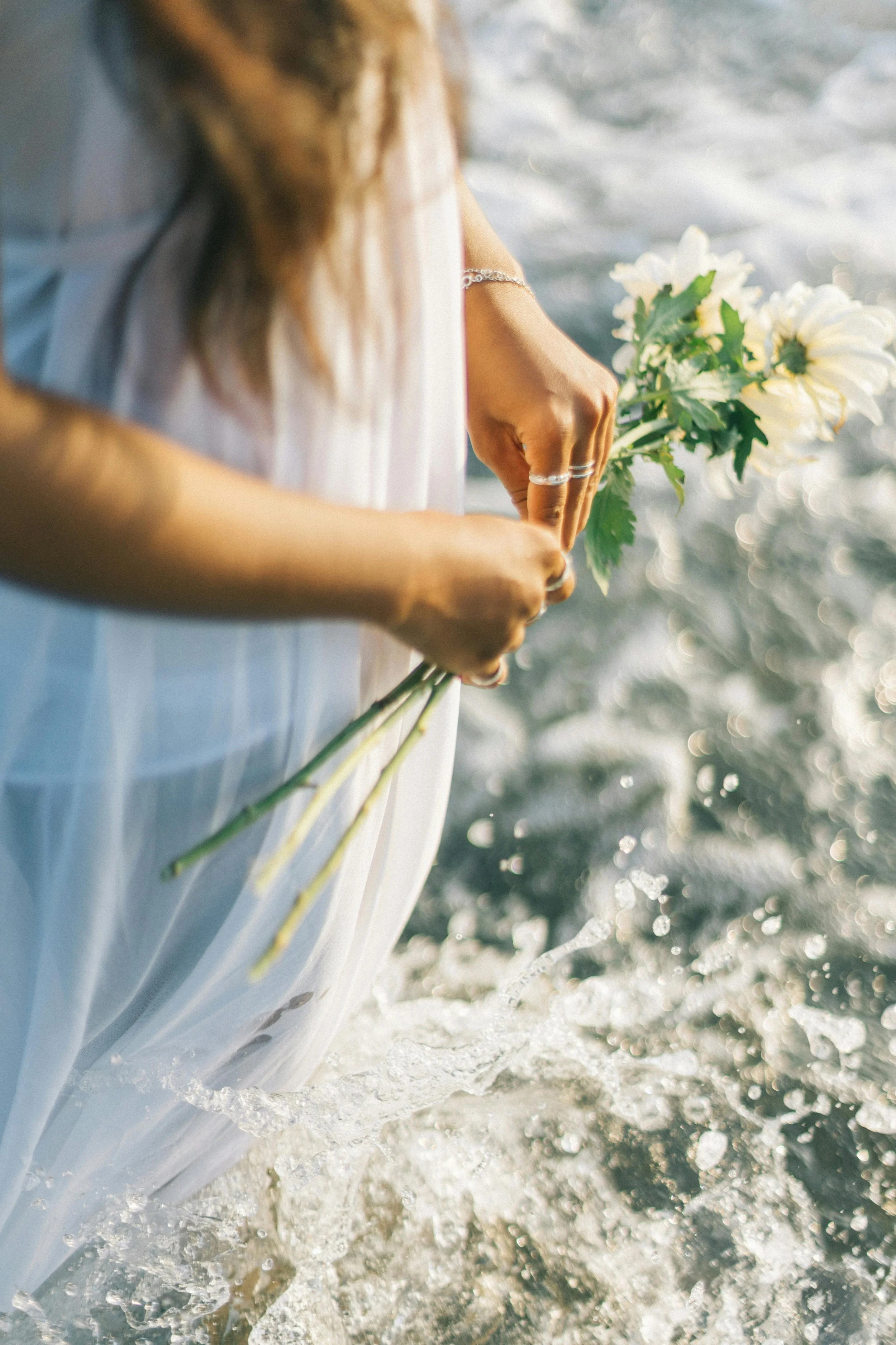 A peaceful, romantic view of a woman holding flowers by the ocean, with waves gently splashing.
