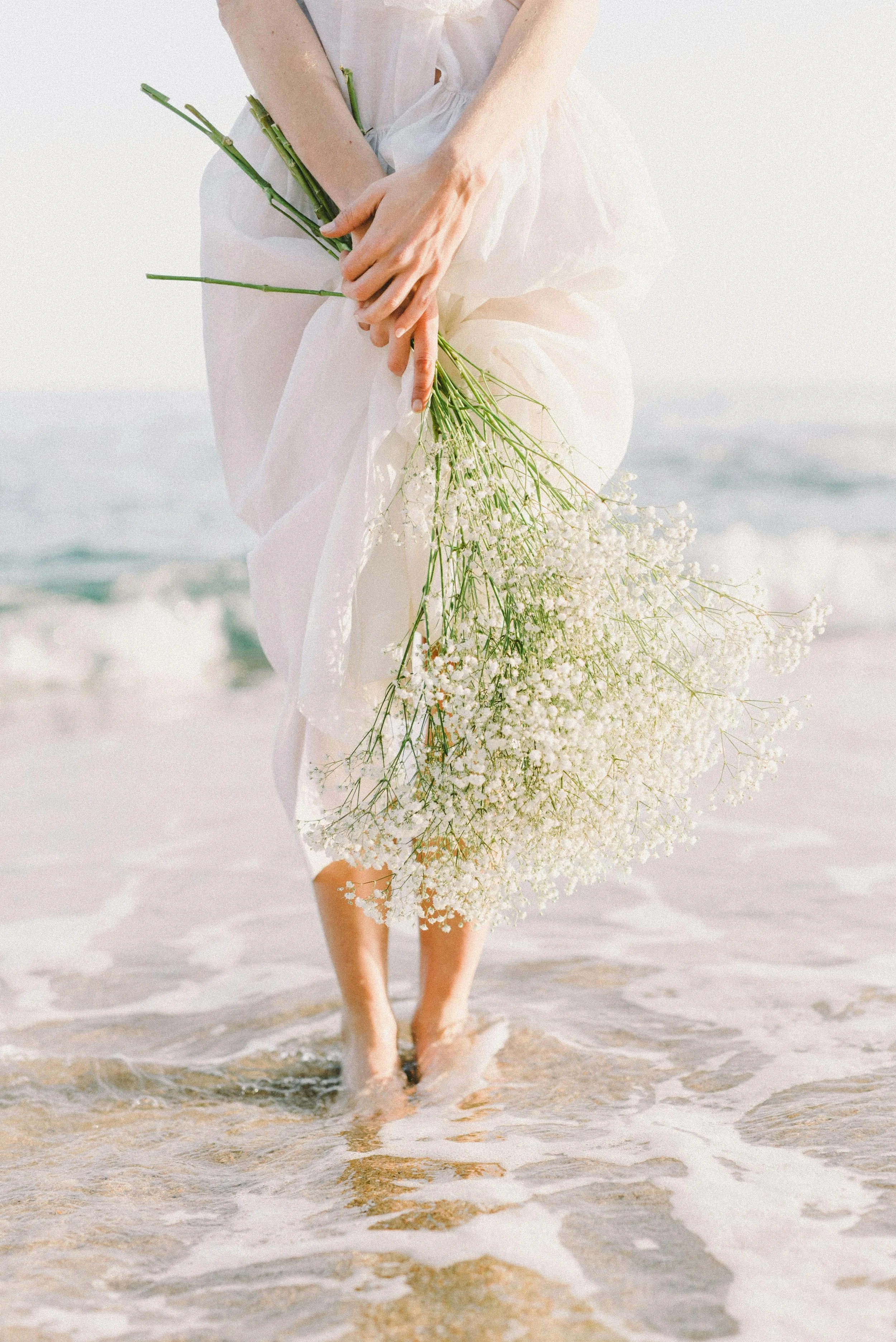 A woman in a white dress holding a flower while walks barefoot along a sunlit beach with gentle ocean waves.