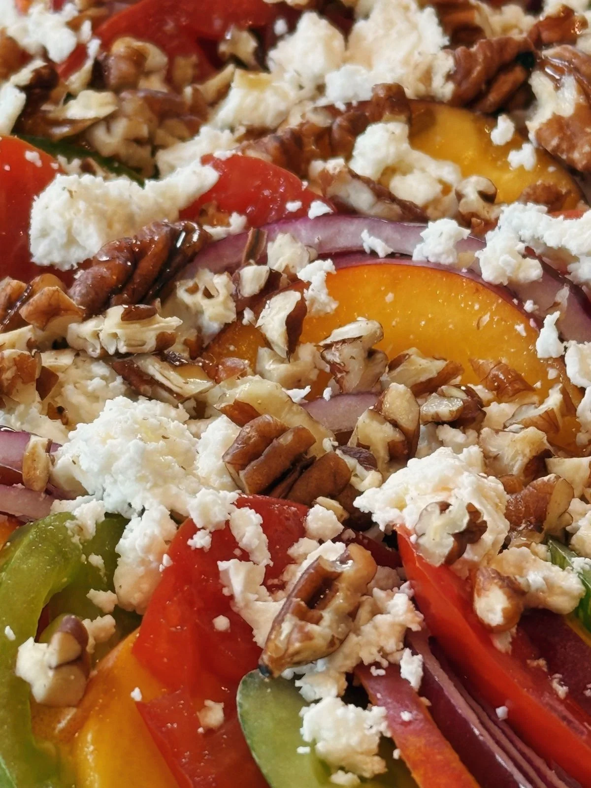 Close-up of a colorful salad with sliced tomatoes, yellow and green bell peppers, chopped onions, crumbled white cheese, chopped nuts, and a drizzle of dressing.
