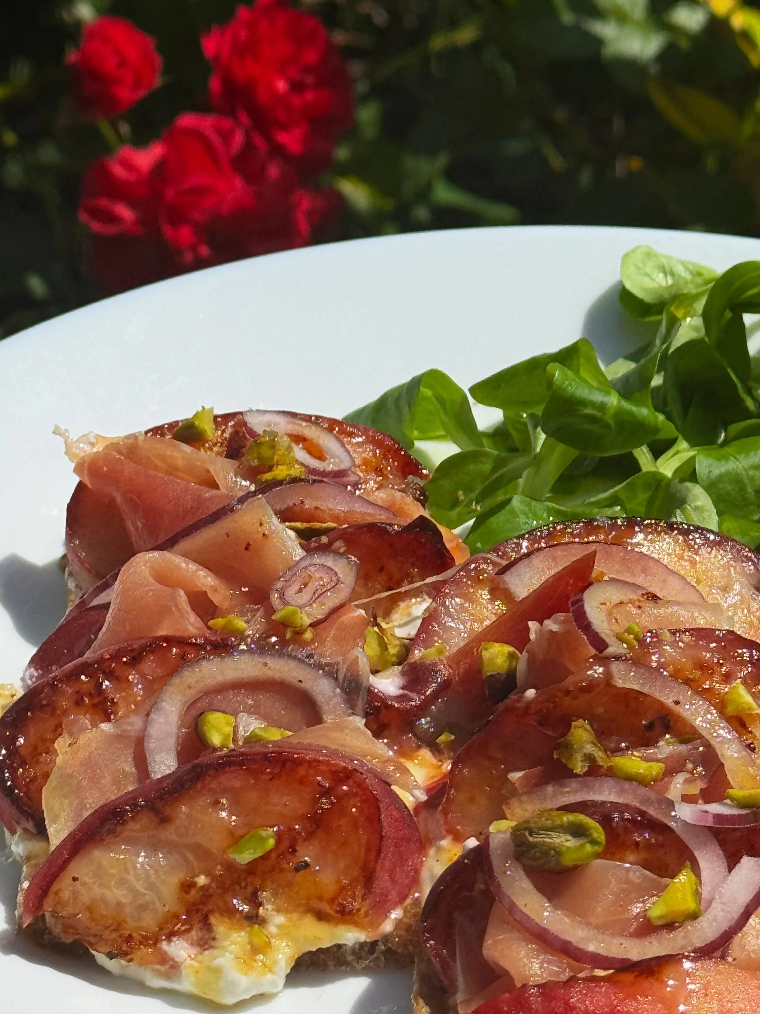 Close-up of a white plate with sliced tomatoes, onions, and green herbs, with red flowers and green leaves in the blurred background.