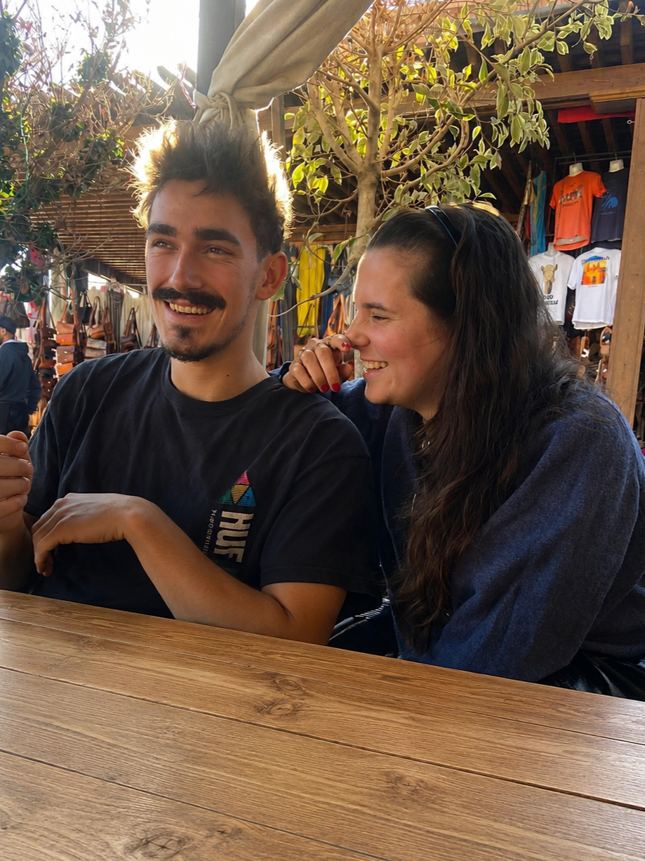 A young man and woman sitting at a wooden table outside, smiling and laughing together with a market or mall in the background, surrounded by trees and merchandise.