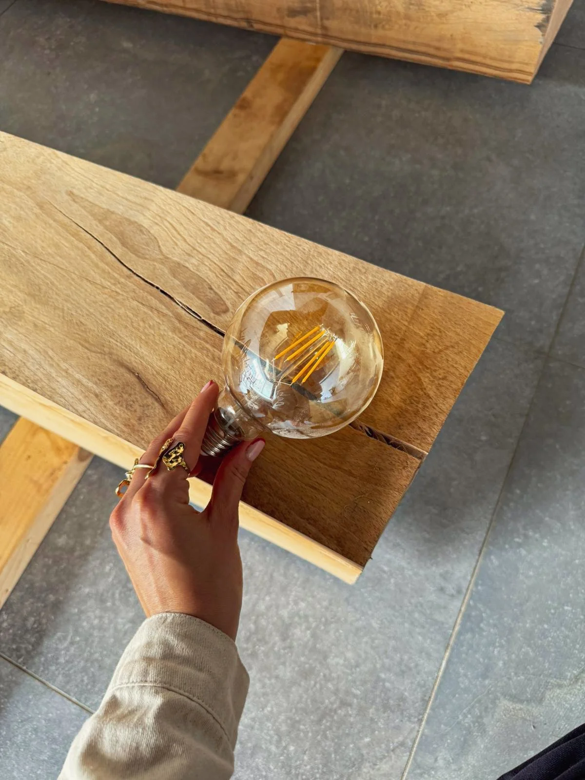 Hand holding a glass light bulb with a visible filament, resting on a wooden surface.