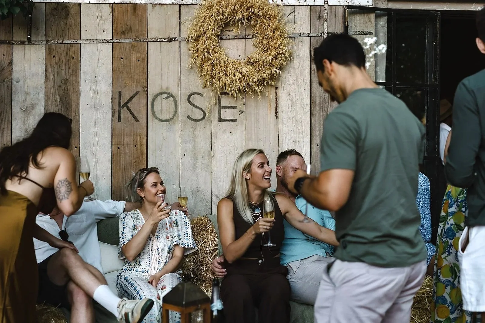 People sitting on hay bales at a rustic indoor party, holding drinks and smiling, with a wooden wall and a wreath hanging above that spells 'KOSE'