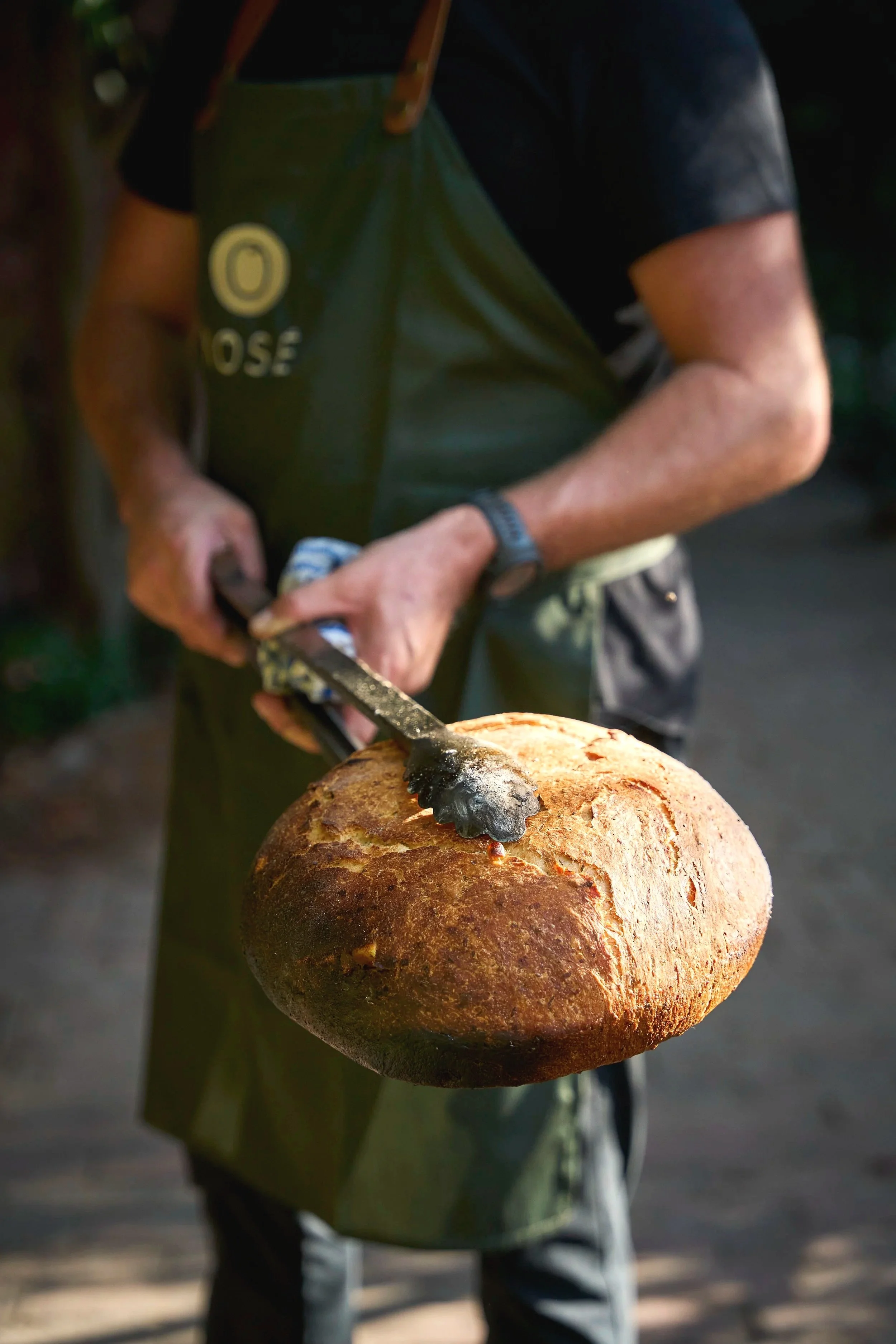 Person holding a freshly baked round loaf of bread with tongs, wearing a green apron and black shirt, outdoors.