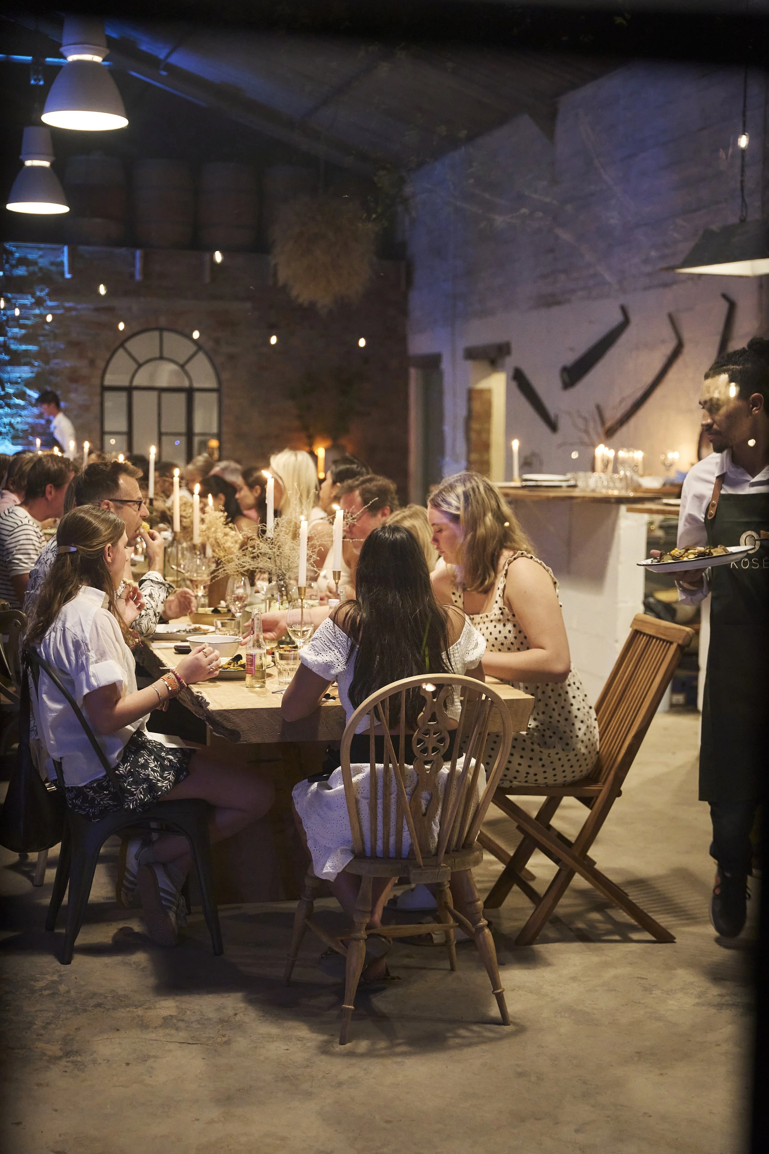 People dining at a long wooden table in a warmly lit restaurant with candles and rustic decor.