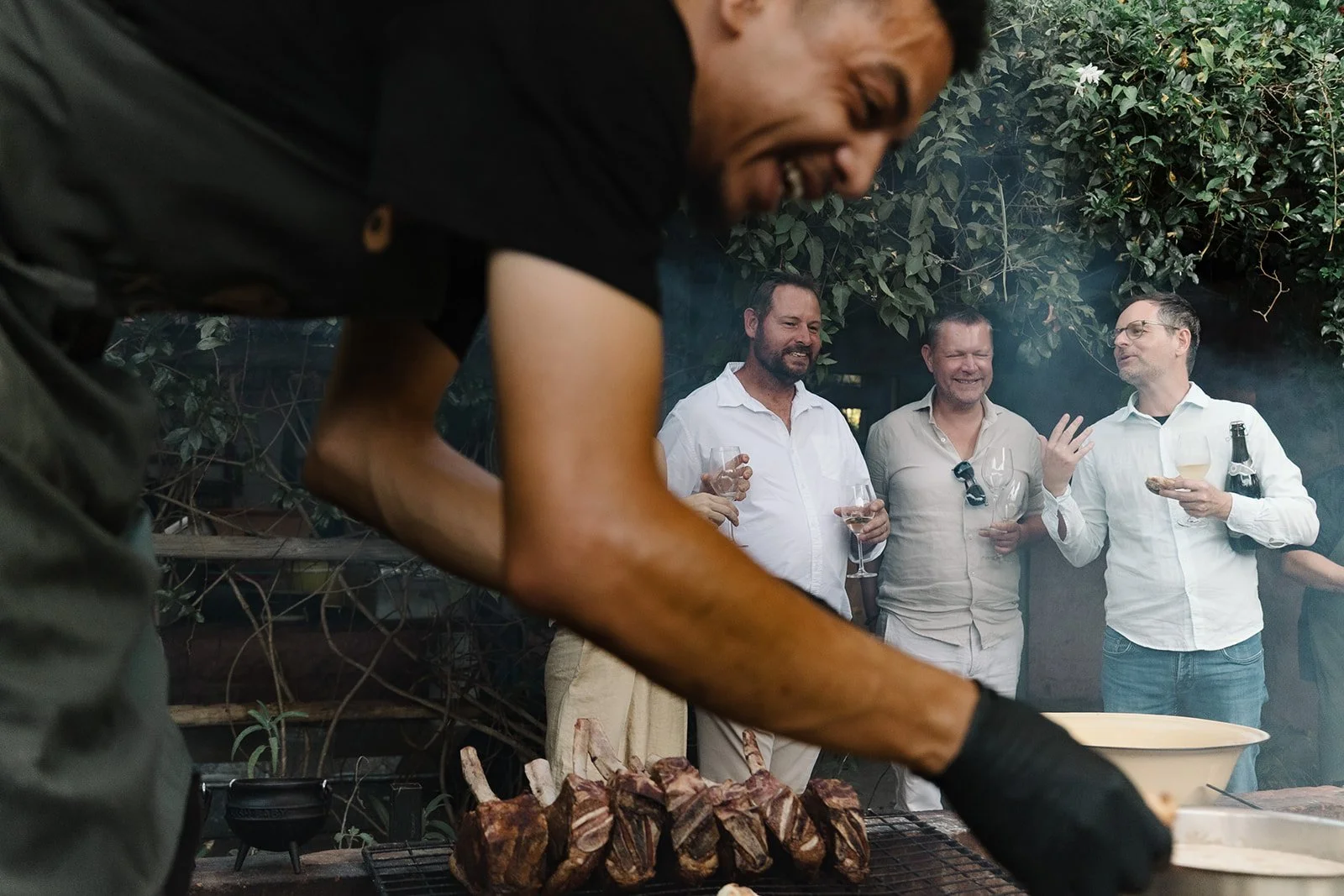 A man is grilling meat, while in the background four men are socializing with drinks in an outdoor setting with greenery.