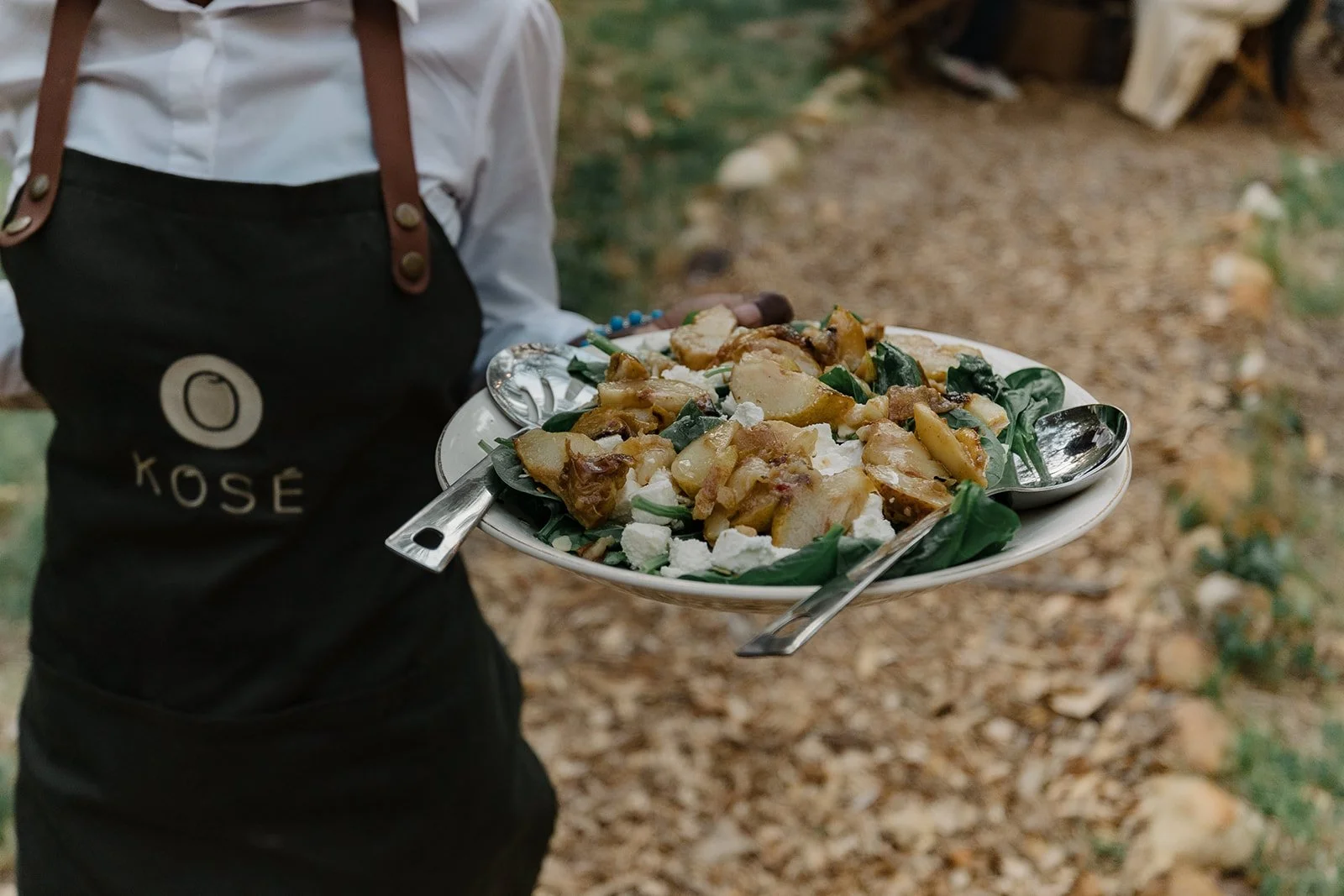 A person wearing a black apron labeled 'Kose' holds a white plate with roasted potatoes, greens, and crumbled cheese, with two spoons on the plate, outdoors on a rocky and leafy ground.