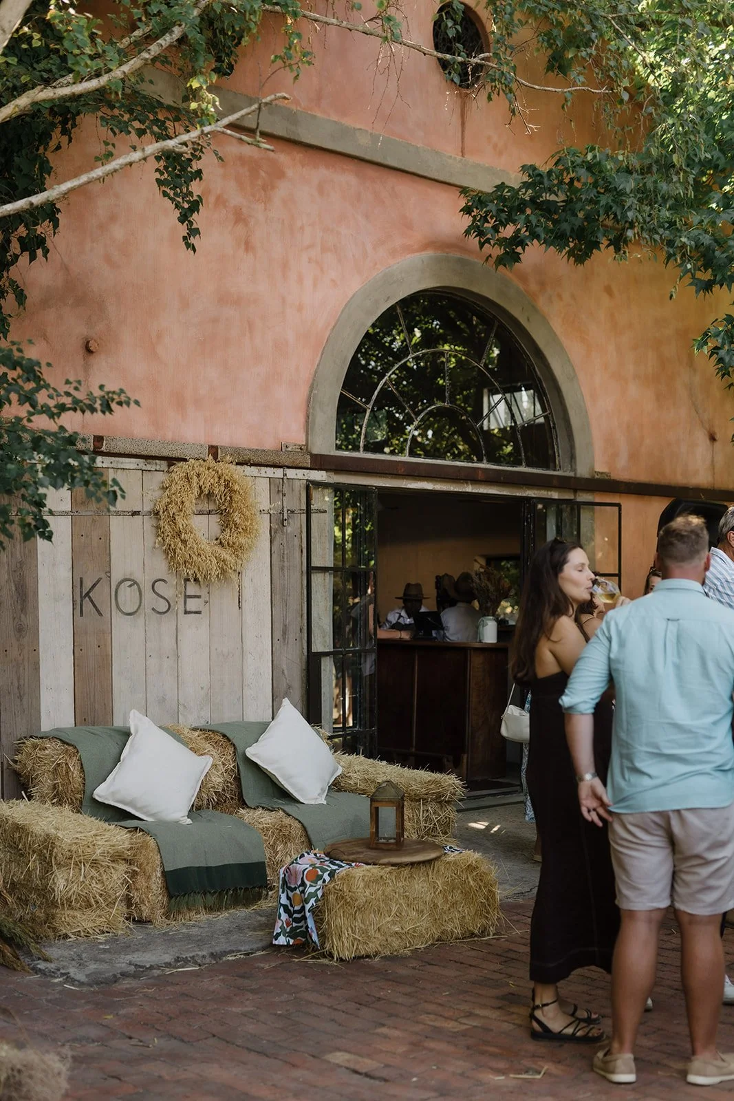 An outdoor gathering area with hay bales used as furniture, decorated with white pillows and a small wooden table, set against a rustic wall with a wreath. People are socializing near an open entrance to a building with a large arched window, under a