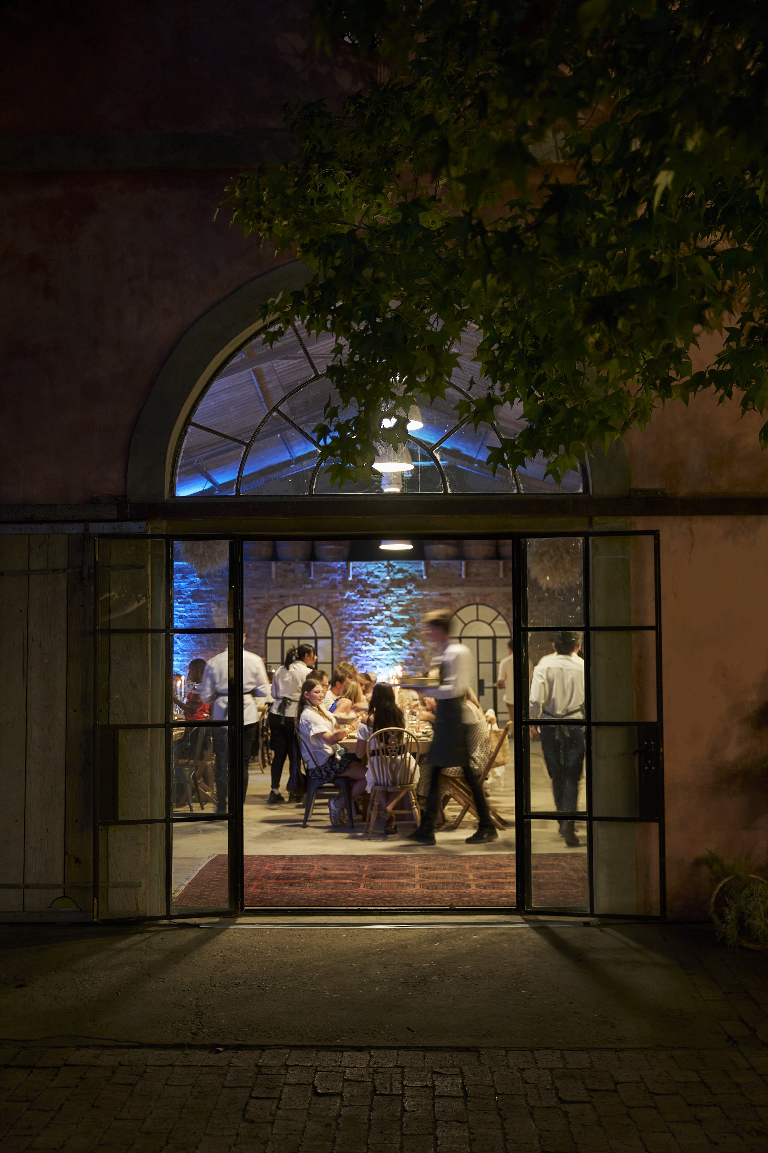 Night view of a restaurant with large glass doors open to reveal a lively dining scene inside, with waitstaff attending to customers and exposed brick walls illuminated by blue accent lighting. A tree partially obscures the top of the restaurant's ar