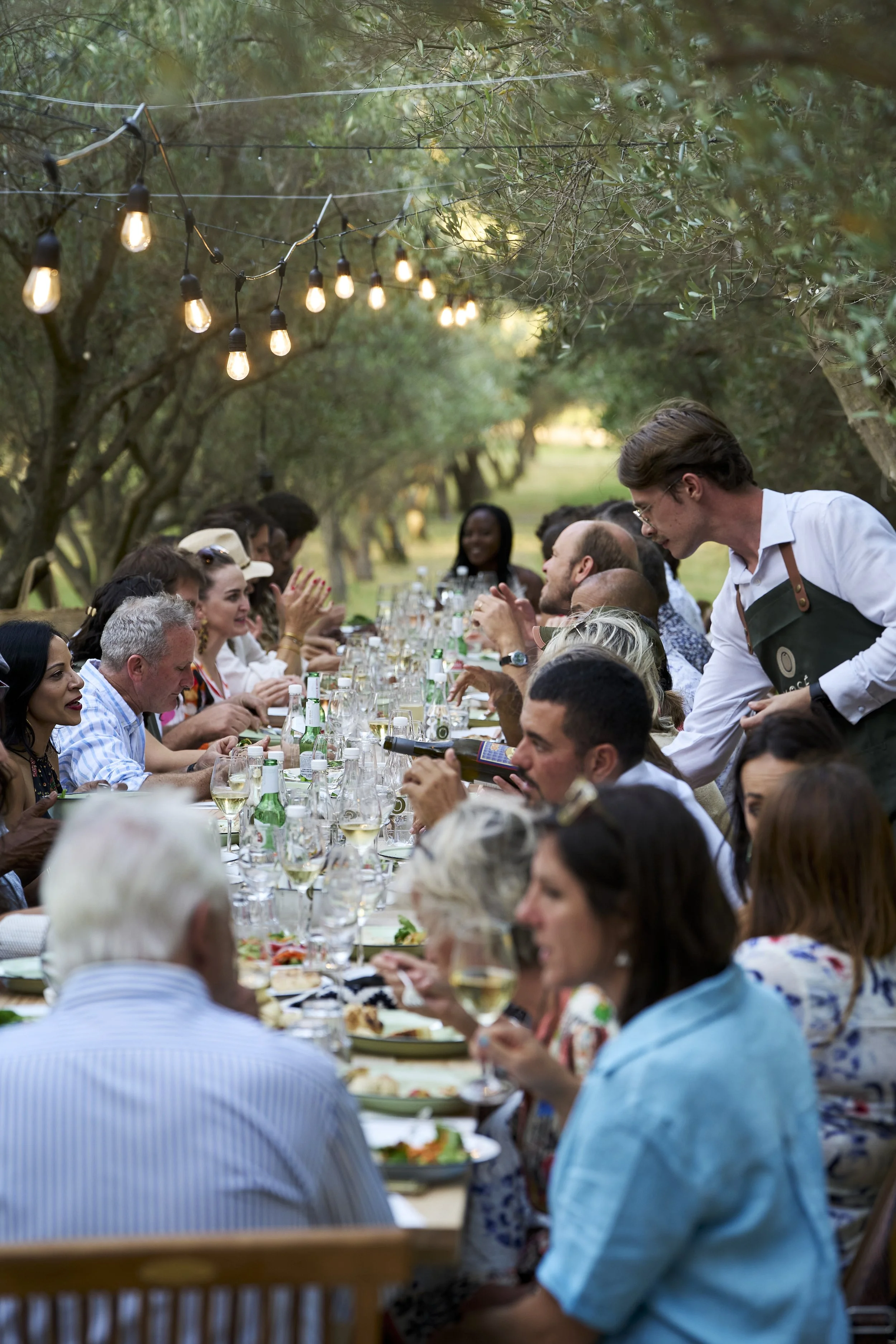 People dining outdoors at a long banquet table with string lights hanging above in a wooded area during evening.