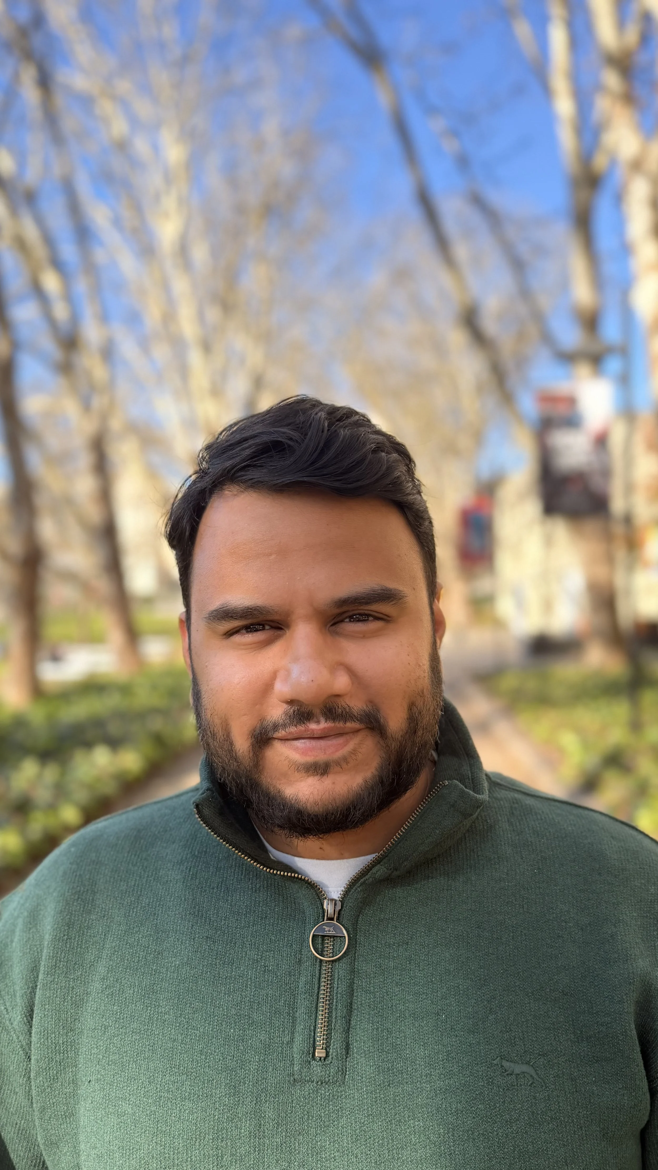 A man with dark hair and a beard, wearing a green zip-up sweater, smiling outdoors with trees and a blue sky in the background.