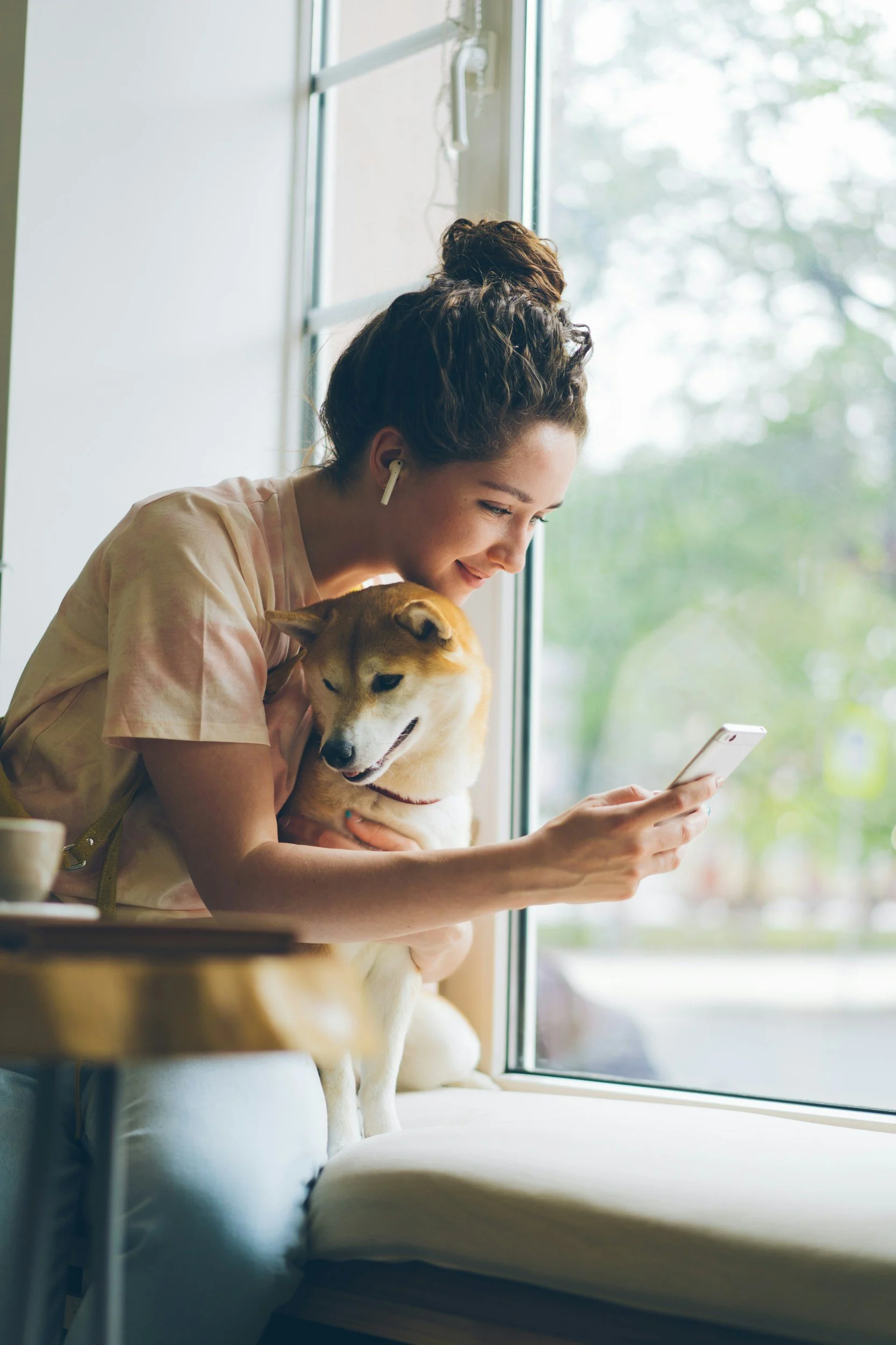 En kvinne med brunt hår i en opphøyet eller knute, smilende og holder en hund på fanget, ser på telefonen sin. De sitter ved et vindu som har et utendørs bakgrunn av grønne trær.