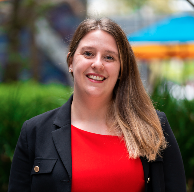 A woman smiling outdoors in front of green plants and a blue umbrella, wearing a red top and a black blazer.