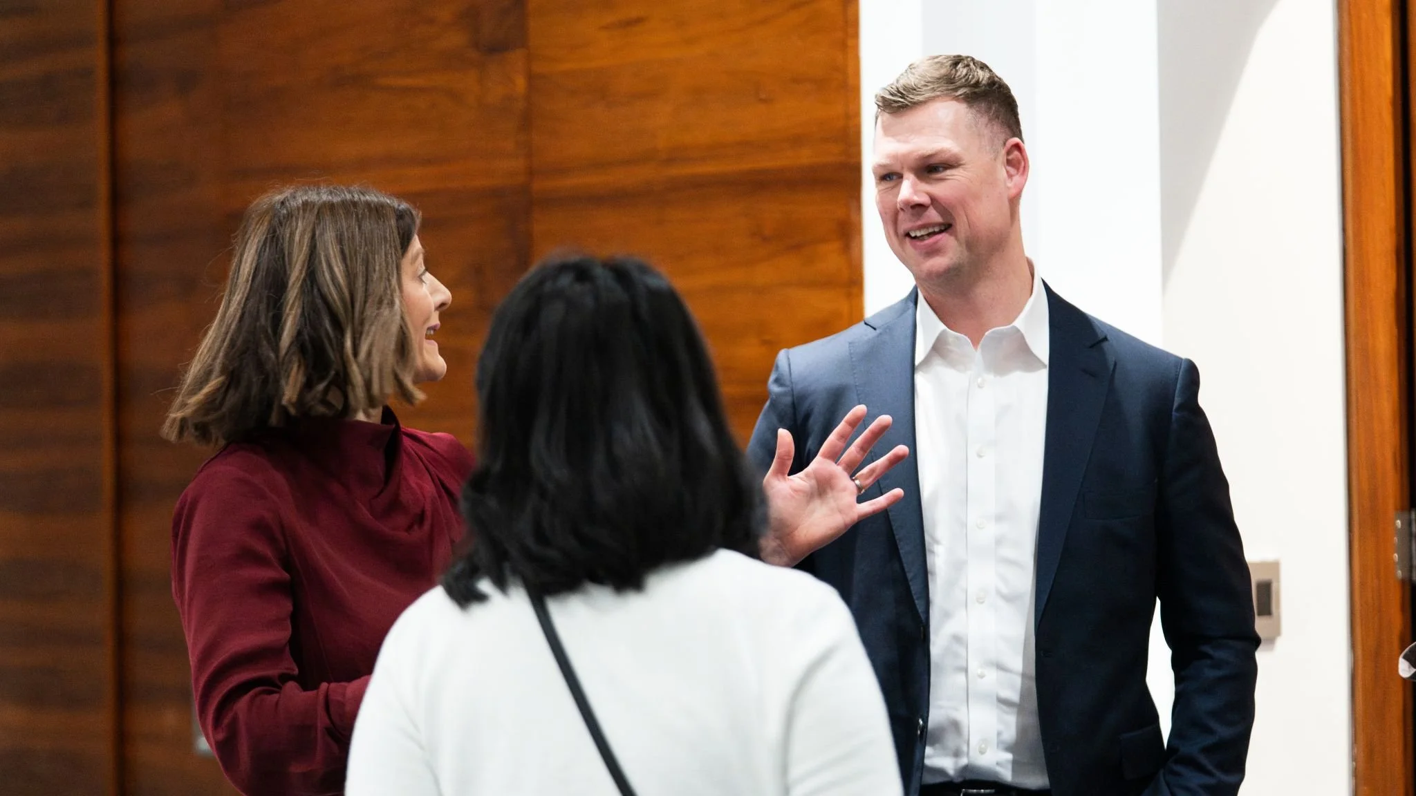 Three professionally dressed people having a conversation in a well-lit room with wooden paneling. The woman on the left, wearing a burgundy top, appears to be speaking, while the man on the right in a navy blazer and white shirt is smiling and gesture with his hand. The woman in the center with black hair, seen from the back, is listening.