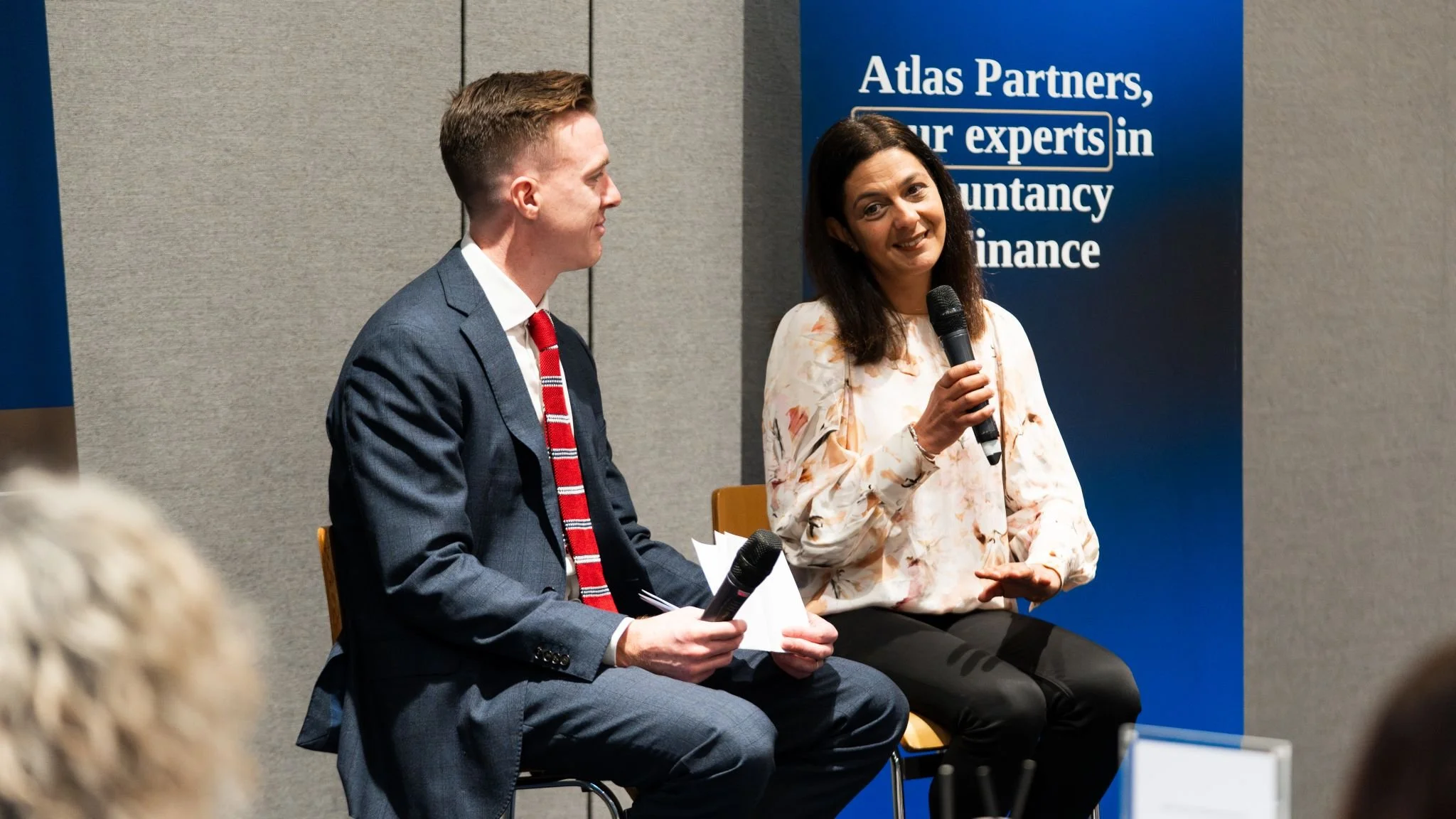 A woman holding a microphone speaking on a panel with a man in a suit holding notes and a microphone, against a blue banner that reads 'Atlas Partners, our experts in accountancy and finance'.