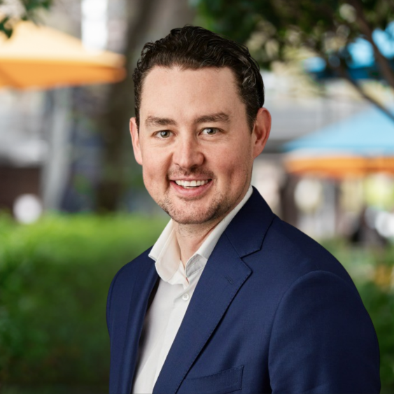 A smiling man in a navy blue suit and white shirt outdoors with blurred umbrellas and greenery in the background.