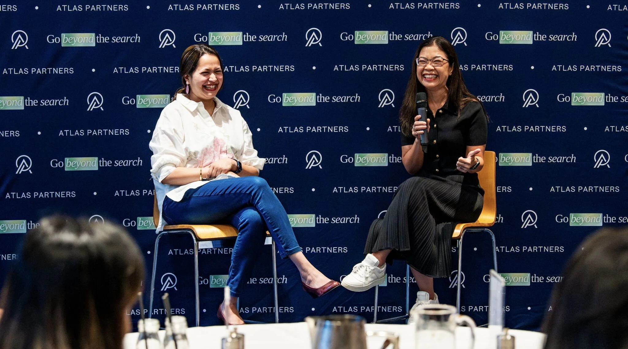 Two women sitting on stage chairs, engaging in conversation with one holding a microphone, set against a blue backdrop with ATLAS PARTNERS and 'Go beyond the search' written repeatedly.