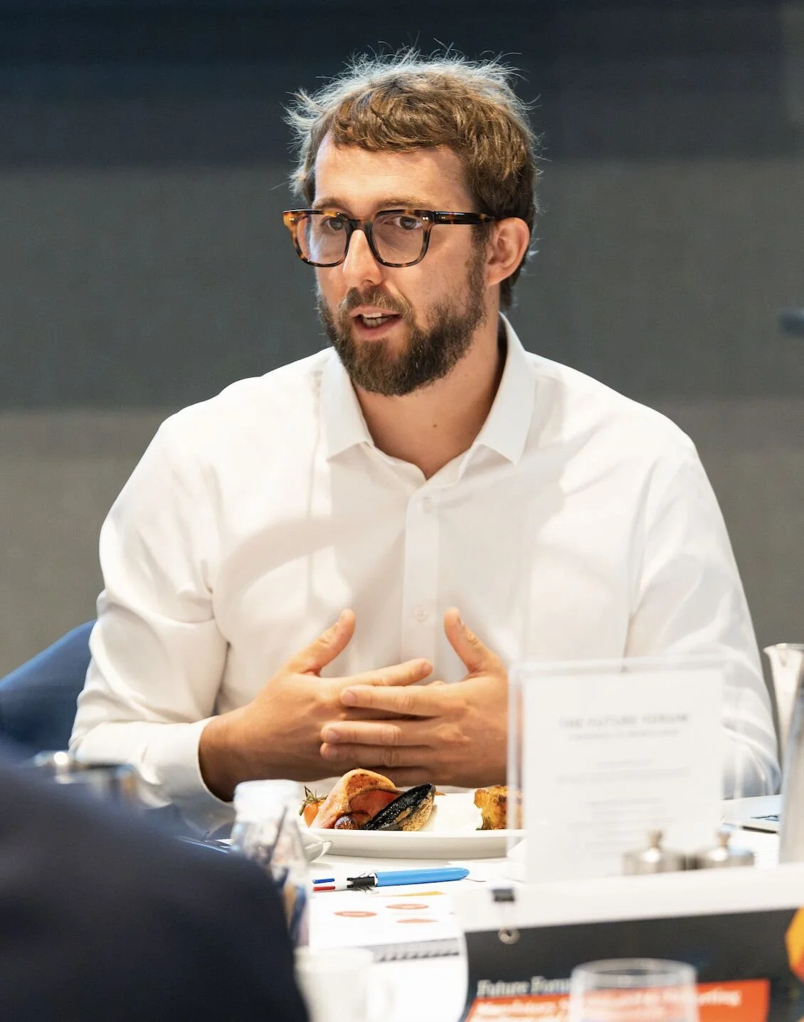 Man with glasses and beard wearing a white shirt, speaking at a meeting.