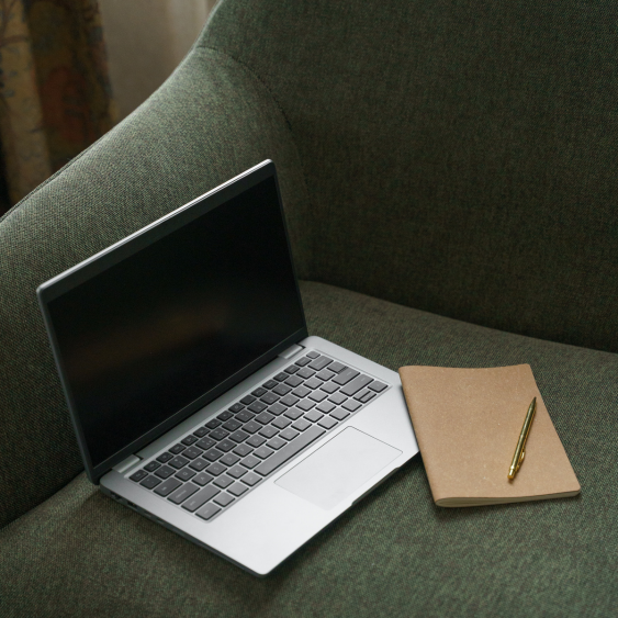 Open silver laptop with black screen next to a closed brown notebook and gold pen on a green sofa.
