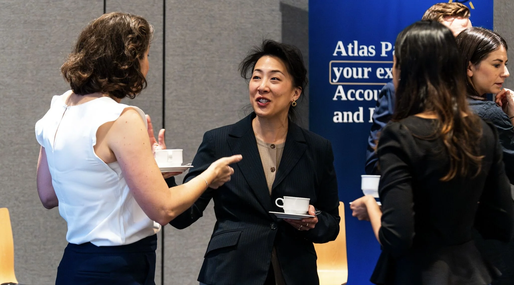 Group of people in a business setting engaging in conversation, with some holding cups and saucers, and a large blue banner in the background.