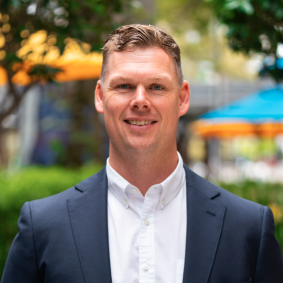 A smiling man in a white shirt and navy blazer outdoors with blurred trees and umbrellas in the background.
