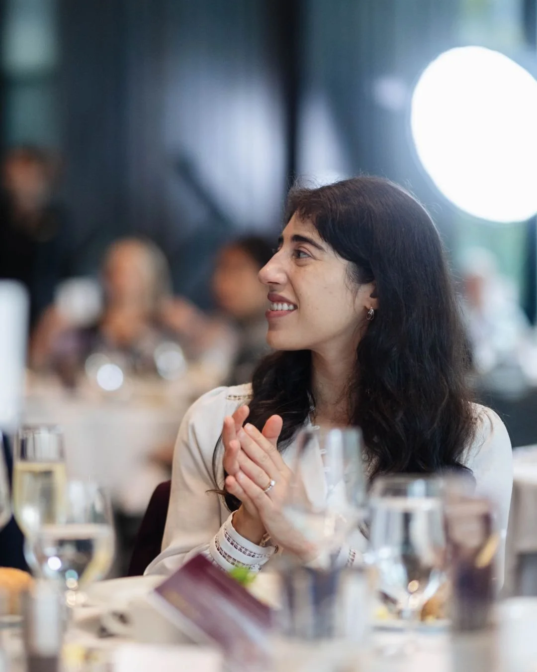 A woman with dark, wavy hair smiling and clapping at a formal dining event, with glasses and plates on the table.