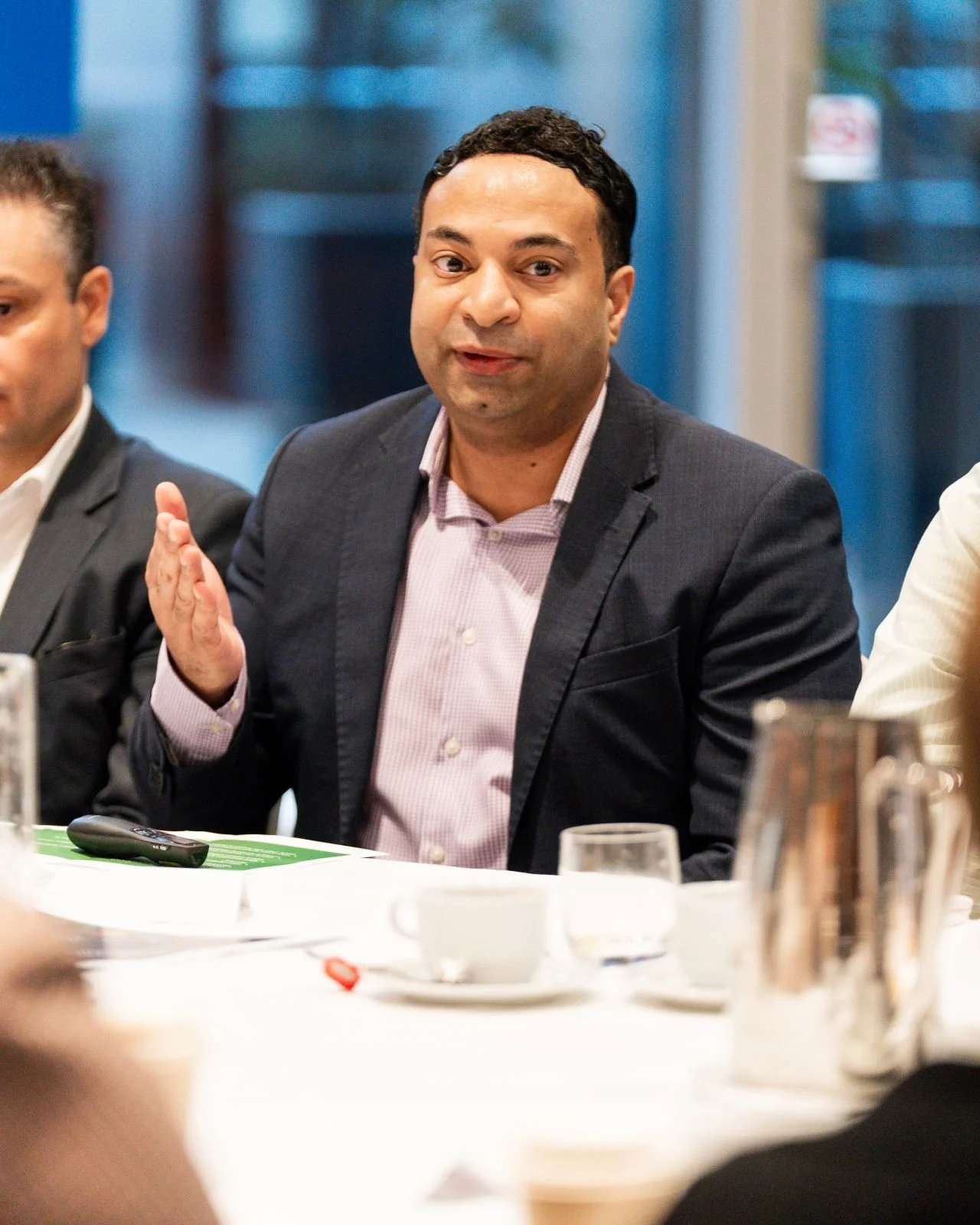 A man in a dark suit speaking at a meeting, with a serious expression, seated at a table with coffee cups, glasses, and papers.