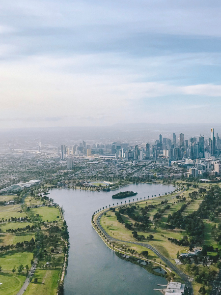 Aerial view of a city skyline with a river and green park in the foreground under a partly cloudy sky.