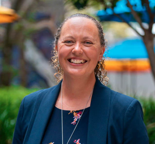 Smiling woman with curly hair wearing a navy blazer and floral top outdoors during daytime