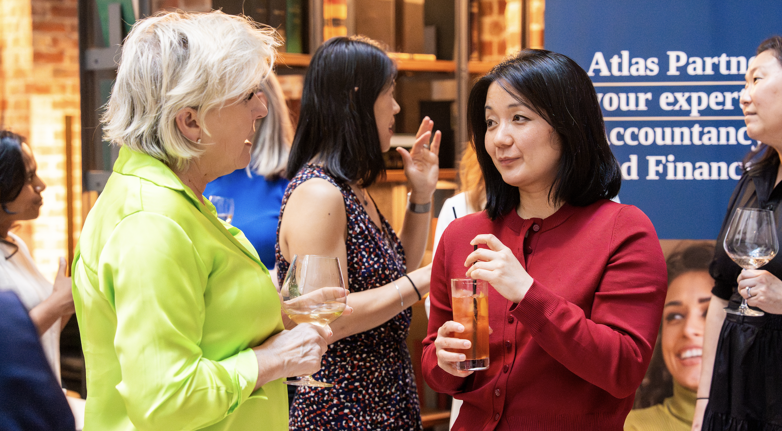 Women at a social gathering, holding wine and drinks, engaging in conversation in an indoor setting with warm lighting and a blue banner in the background.