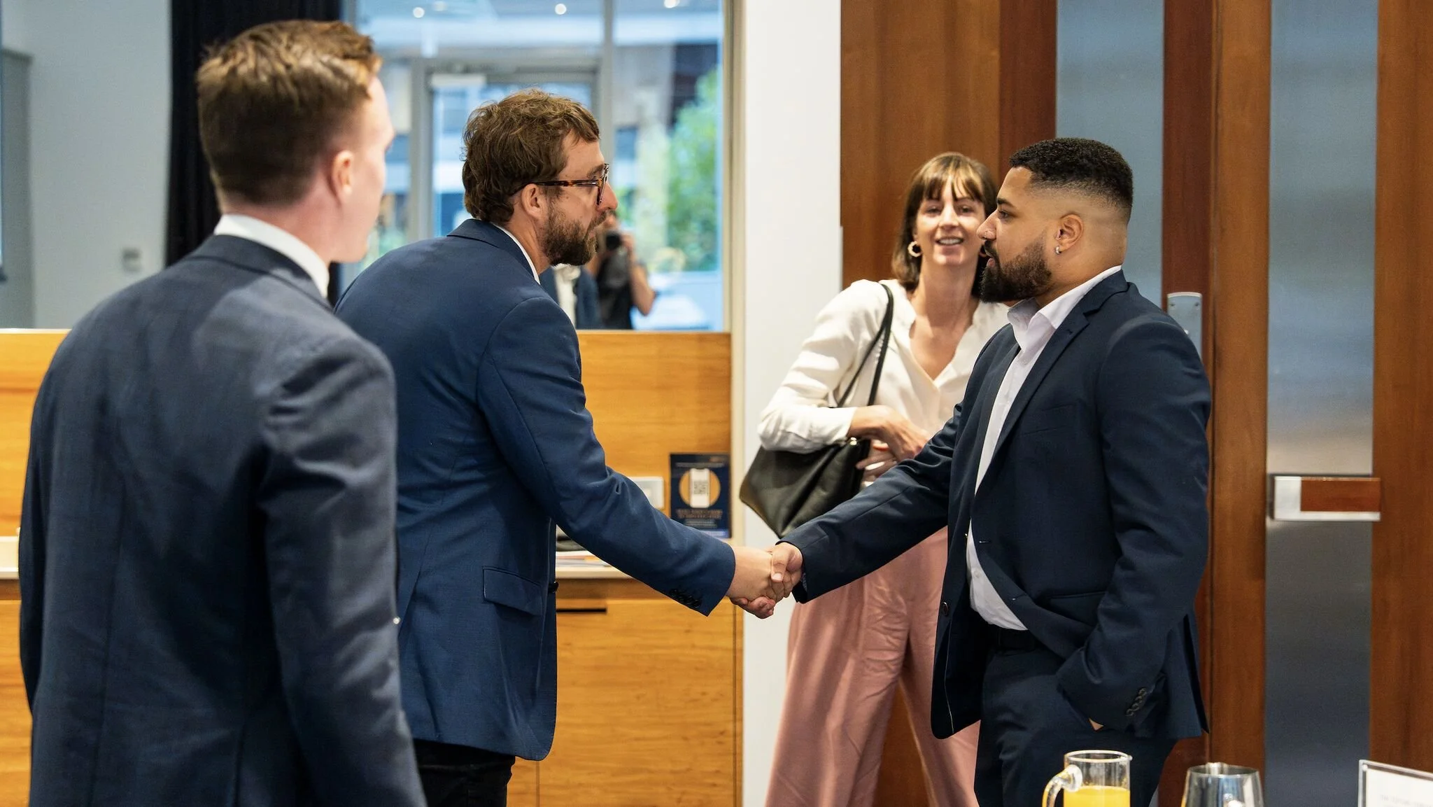 Two men in suits shake hands in a professional setting, with two women in the background observing and smiling.