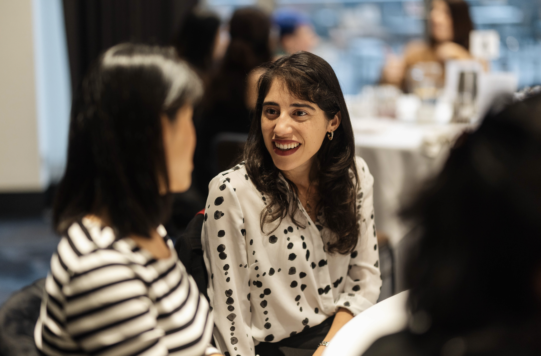 A group of women sitting together at a dining table, engaging in conversation in a social setting, with one woman smiling and looking at another woman.