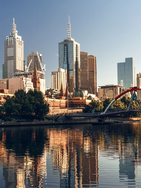 City skyline with tall buildings reflecting in a river, with a bridge and trees in the foreground.