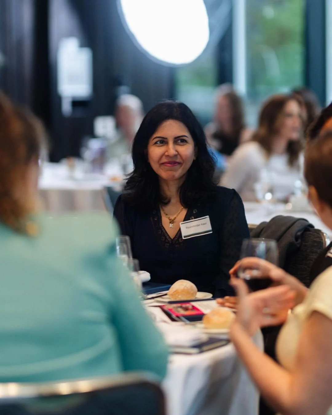 A woman with dark hair and a name tag, sitting at a table during a social event, with other women around her, in a well-lit room with large windows.