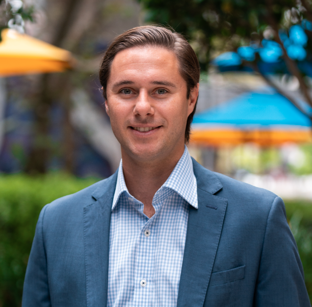 Man smiling outdoors, wearing a blue blazer and patterned shirt, in a park with yellow and blue umbrellas in the background.