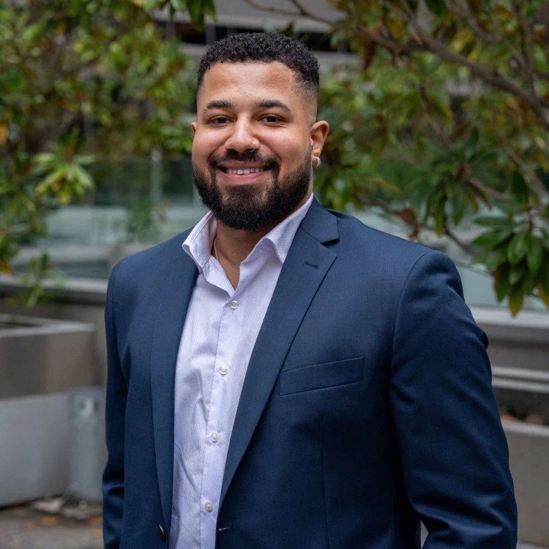 Portrait of a smiling man with a beard in a navy suit and white shirt, standing outdoors with green trees in the background.