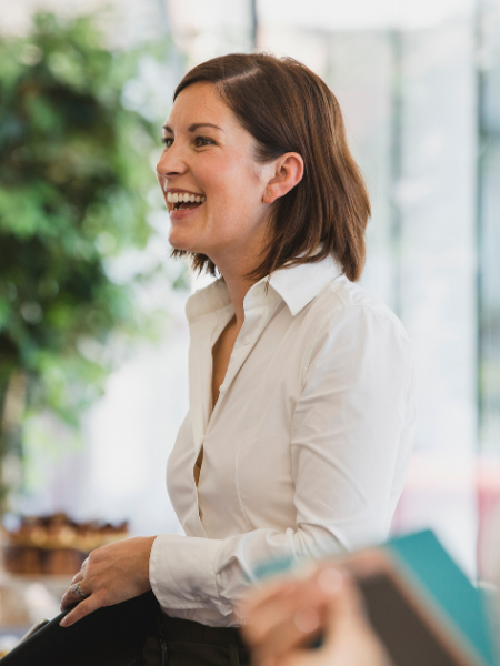 Woman with brown hair smiling and laughing, wearing a white shirt, in a bright indoor setting.