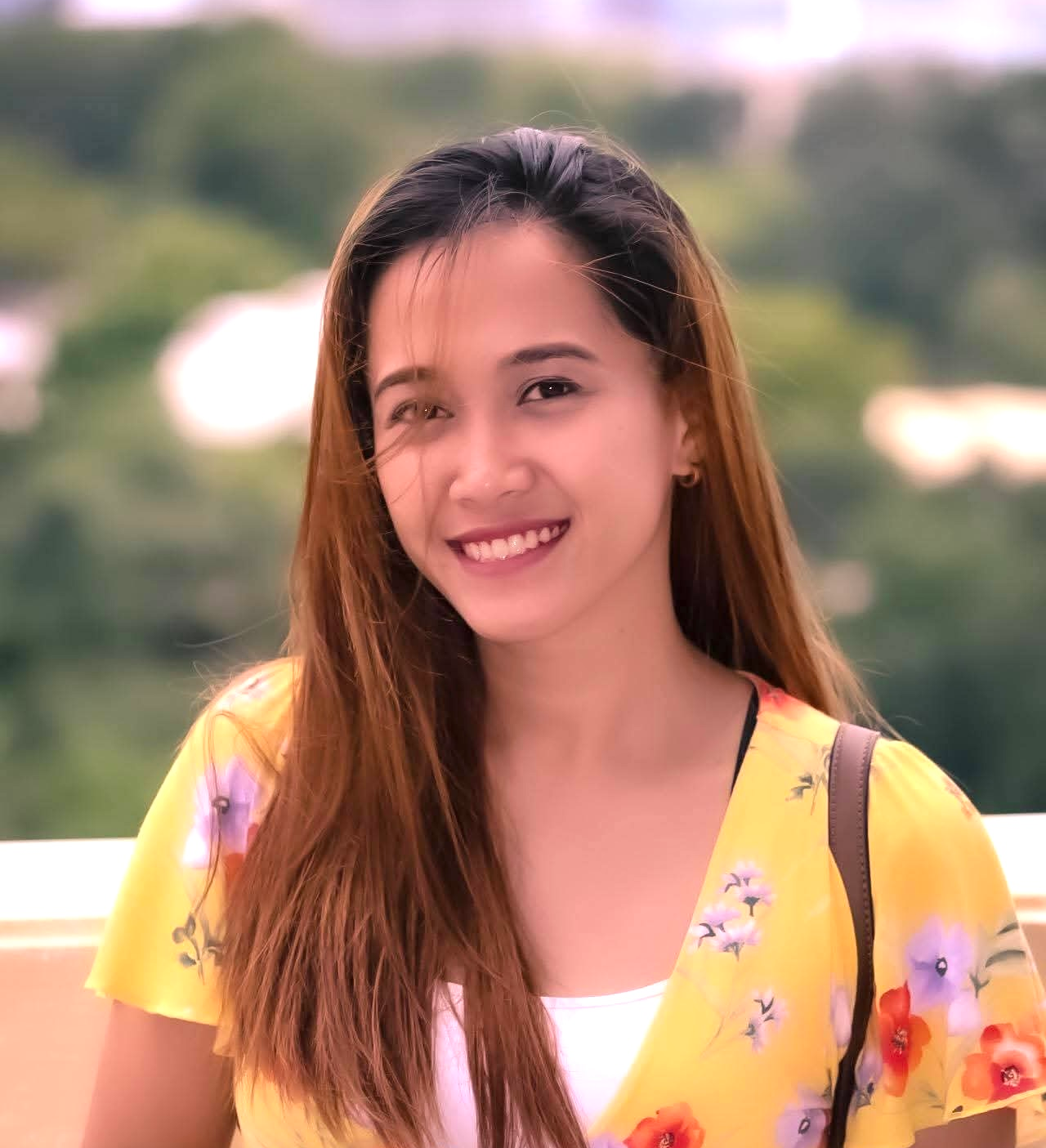 Young woman with long brown hair smiling, wearing a yellow floral dress, outdoors with blurred greenery background.
