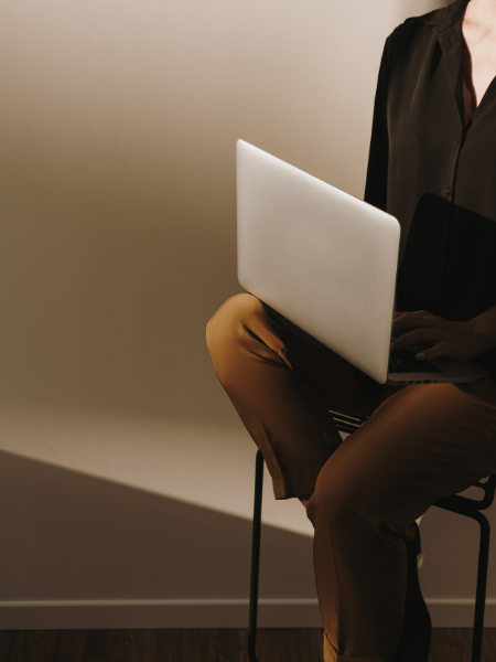 Person sitting on a chair with a laptop on their lap, wearing a black shirt and brown pants, in a plain room.