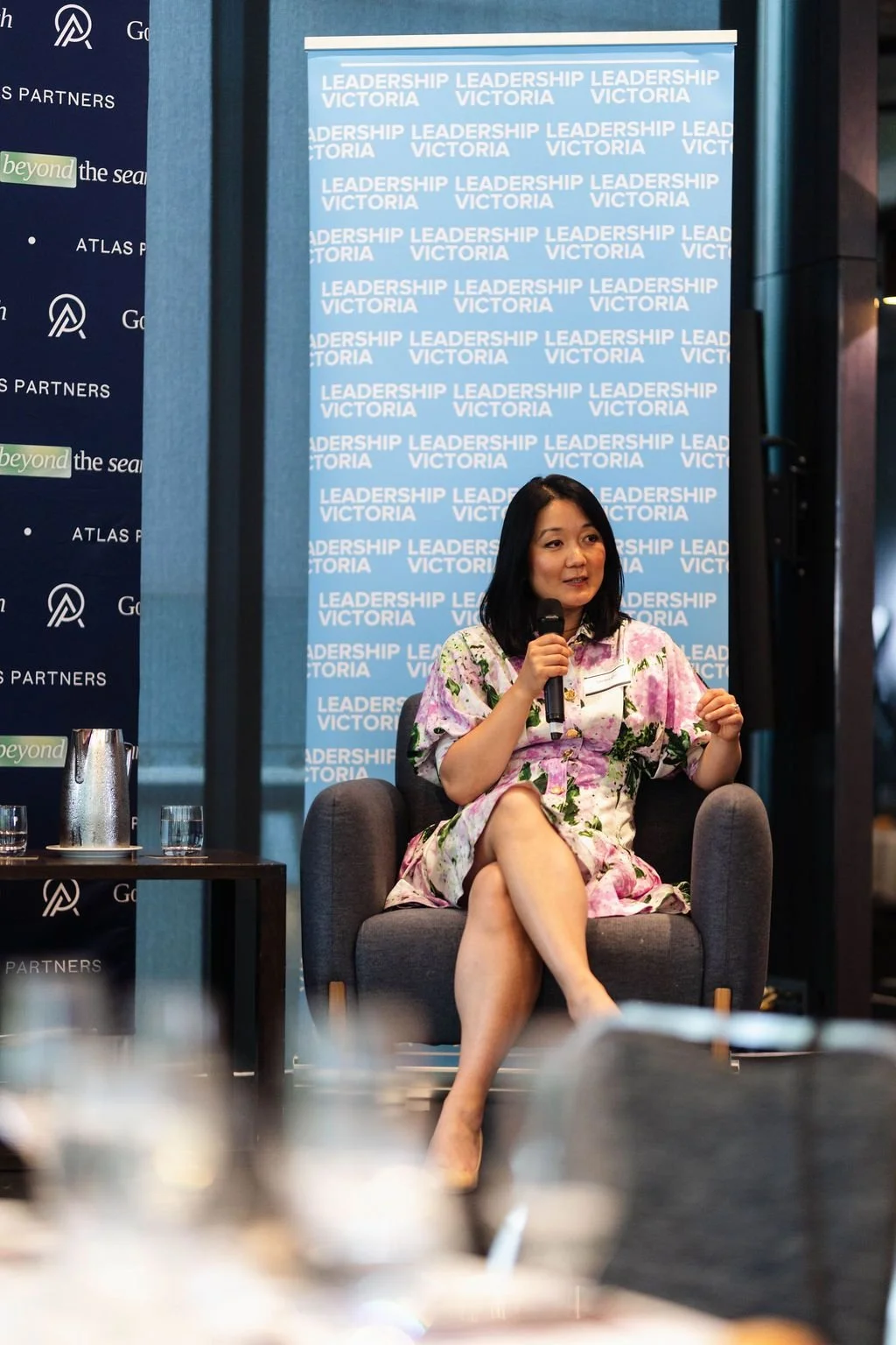 A woman with shoulder-length black hair, wearing a floral dress, is sitting and speaking into a microphone during a panel or presentation at an event. Behind her is a blue backdrop with the words 'Leadership Victoria' repeated.