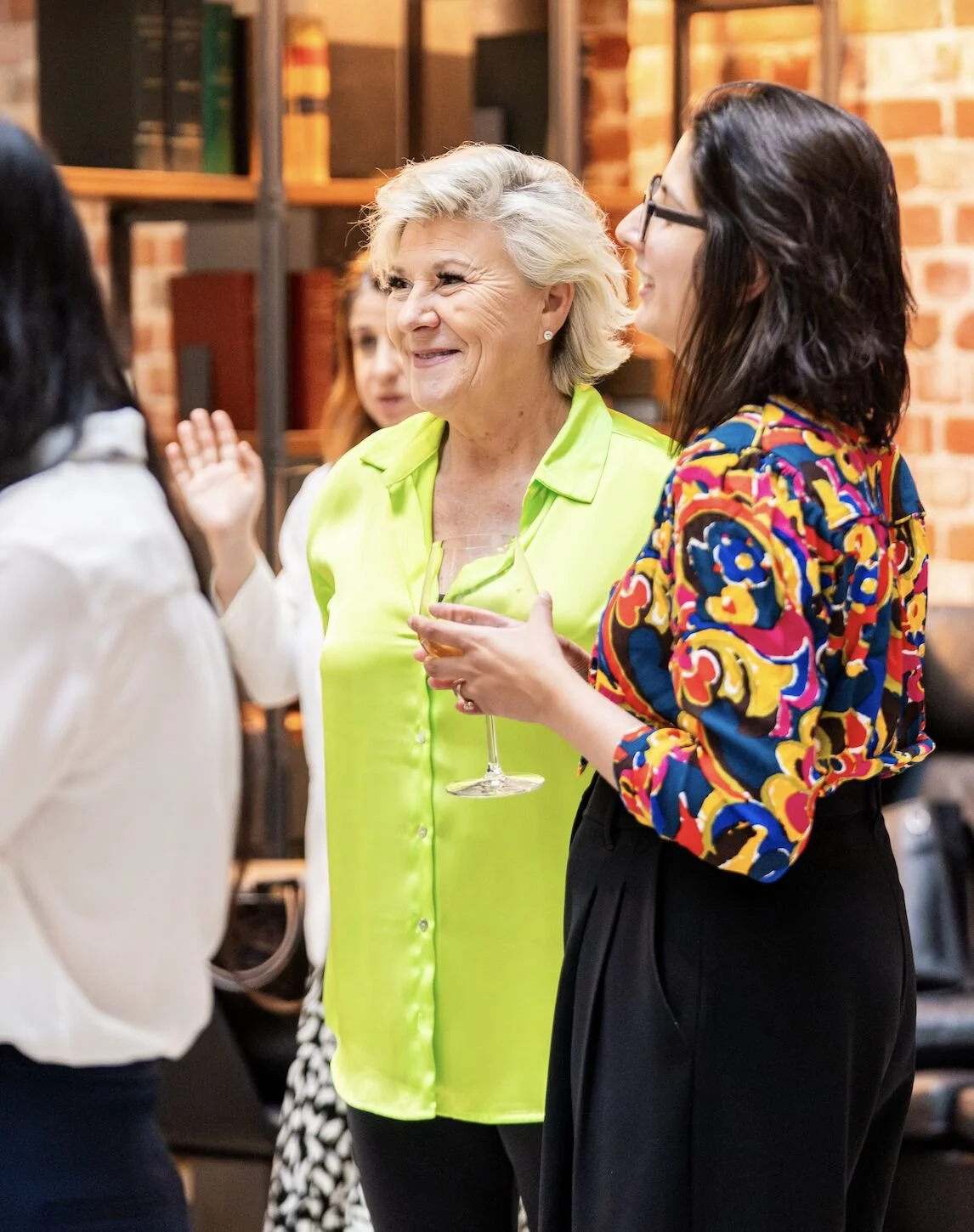 Group of women at a social gathering, talking and smiling indoors with brick walls and shelves in the background.