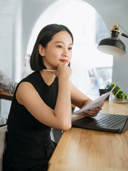 Young woman sitting at a desk, holding papers, looking thoughtfully at a laptop in a modern, well-lit office or cafe.