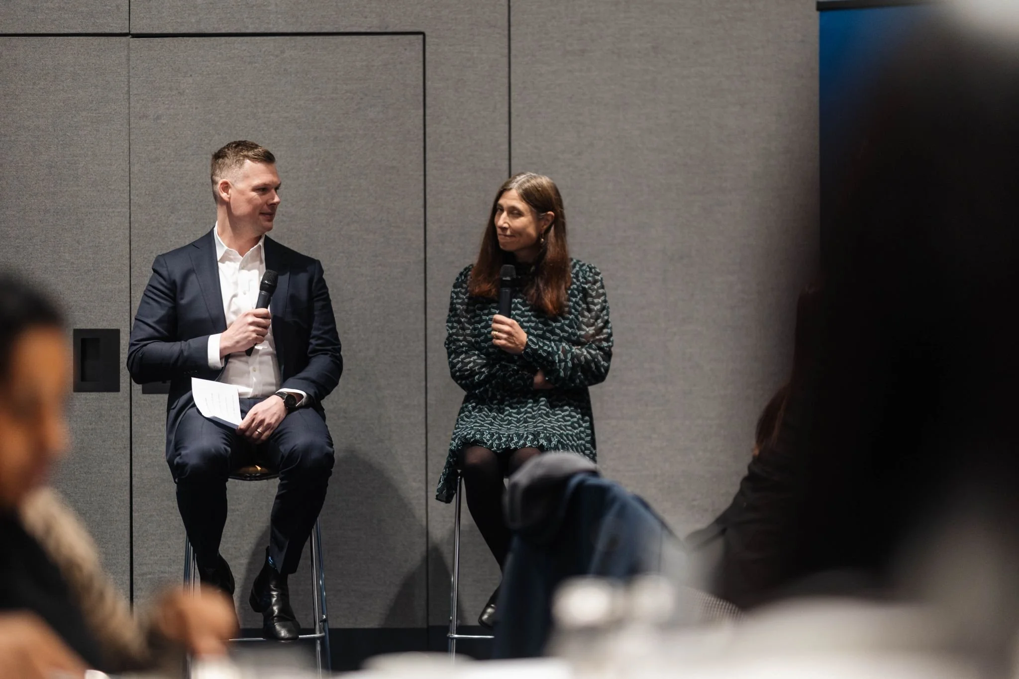 A man and a woman sit on stools on a stage, each holding a microphone, engaging in a discussion or interview during a conference or panel event. The man is wearing a dark suit with a white shirt, and the woman is dressed in a patterned dress with dark tights.