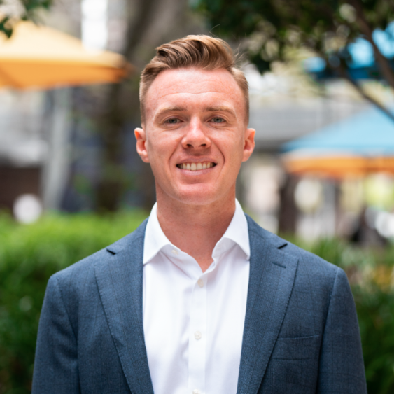 A young man with short, styled light brown hair, wearing a blue suit jacket and white shirt, smiling outdoors with blurred greenery, colorful umbrellas, and buildings in the background.