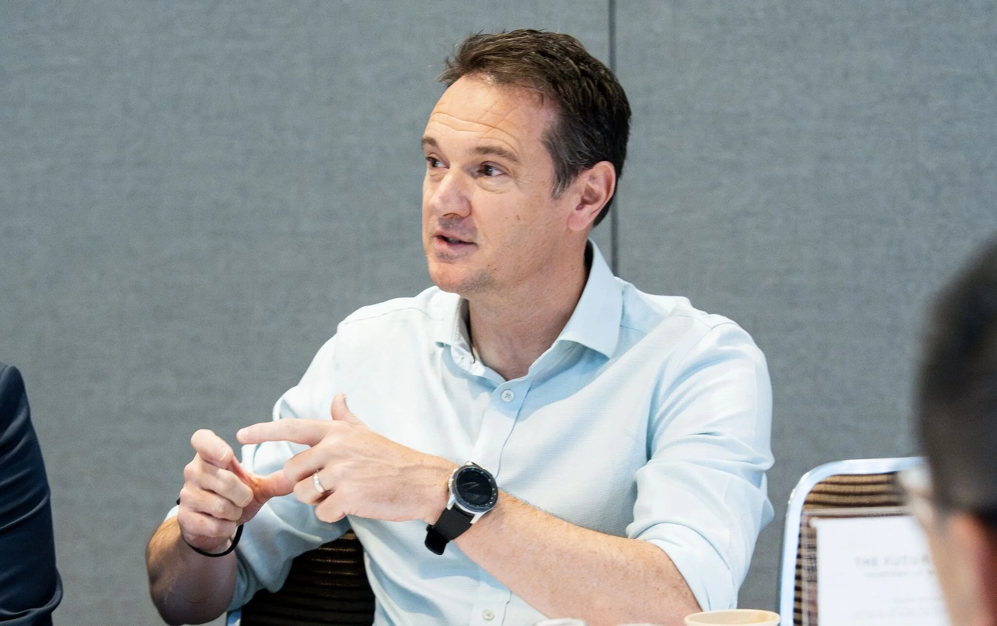 A man sitting at a meeting table, speaking and gesturing with his hands, wearing a light blue shirt and a smartwatch.