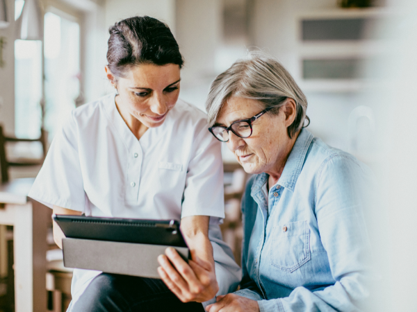 A caregiver showing something on a tablet to an elderly woman in a bright, modern kitchen.