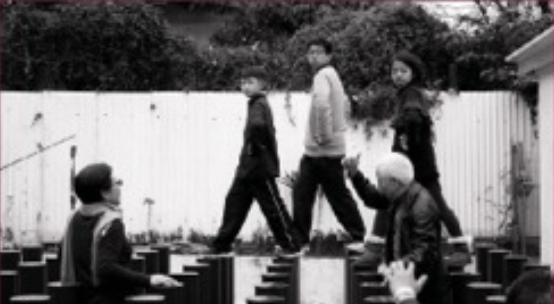 Five children playing outside on large wooden blocks in front of a white fence, with trees in the background.